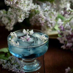 Artistic shot of a blue cocktail adorned with lilac flowers, captured with a dreamy floral backdrop.