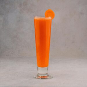 Bright carrot juice served in a tall glass with a slice for garnish on a gray background.
