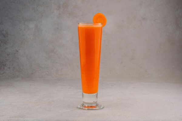 Bright carrot juice served in a tall glass with a slice for garnish on a gray background.