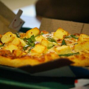 A delicious cheesy pizza topped with paneer and peppers, served in a cardboard box. Mouth-watering and ready to enjoy.