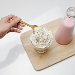 High angle of crop unrecognizable woman checking by wooden teaspoon froth of whipped cream on dessert served near bottle with pink milkshake on wooden board